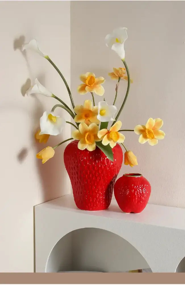Red ceramic flower vase shaped like a cartoon strawberry holding white calla lilies and yellow daffodils for décor