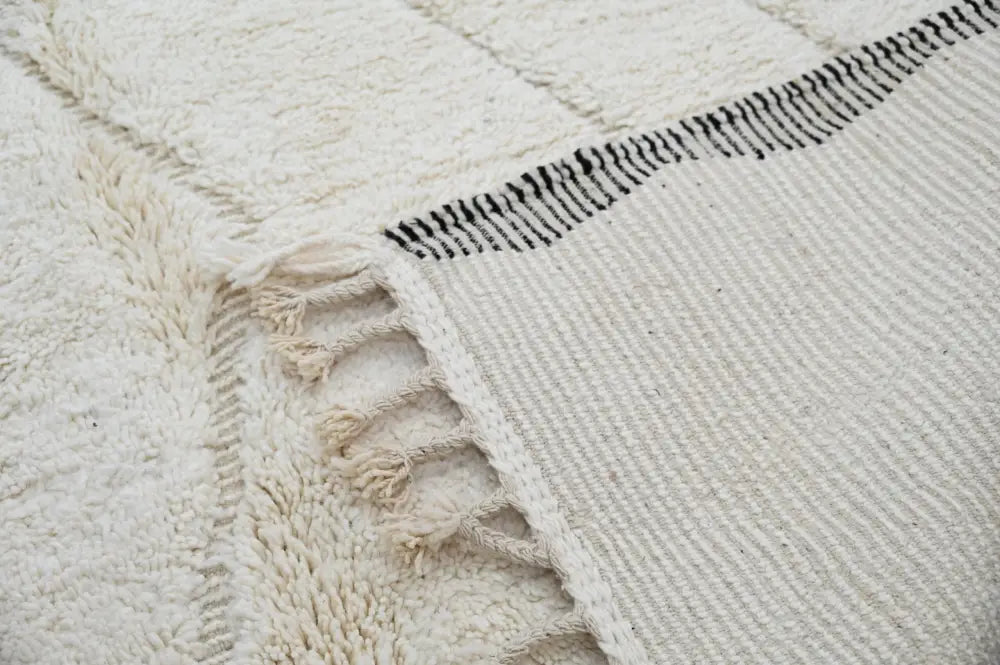 Cream-colored wool tapis beni ouarain with black geometric stitching and fringed edges measuring 290 x 205 cm