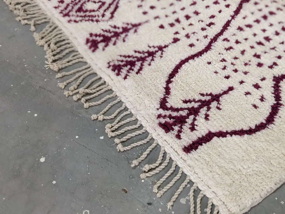A cream-colored tapis beni ouarain with burgundy geometric leaf patterns and a thickly fringed edge by decorbites