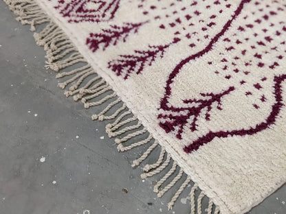 A cream-colored tapis beni ouarain with burgundy geometric leaf patterns and a thickly fringed edge by decorbites
