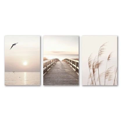 Set of three minimalist white seascape canvas prints featuring a seagull, boardwalk, and tall reeds by DecorBites™