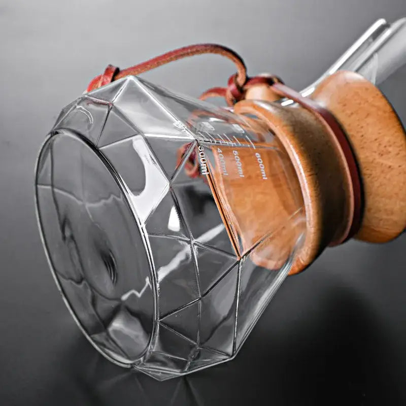 Clear glass hand brew coffee dripper with diamond glass hand design and wooden lid secured by brown leather straps for kitchen use