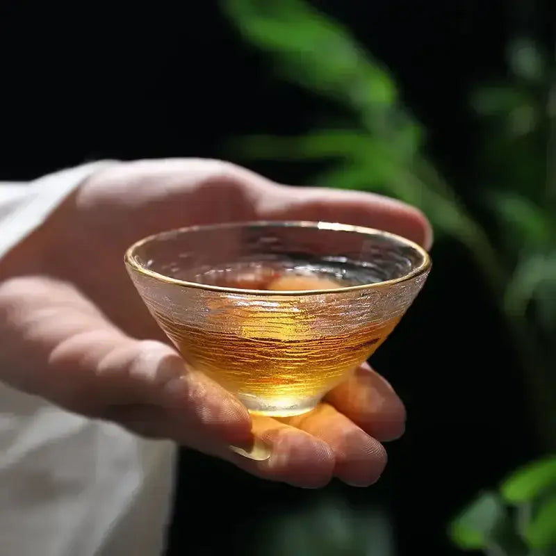 A hand holds an elegant carved tin glass tea cup with a gold rim filled with amber tea from a tea cup set