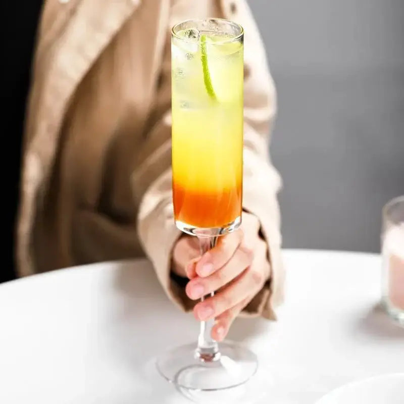 A tall clear glass bubble champagne glass with an orange layered cocktail and lime twist in a decorbites set of 2