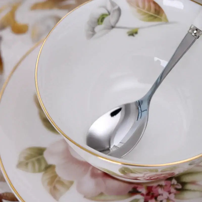 A shiny silver spoon rests inside an elegant floral porcelain tea cup set with gold trim and pink rose design