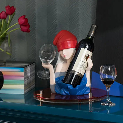 A stylized resin wine rack of an elegant girl sculpture in red beanie and blue dress holding a wine glass and bottle decorbites