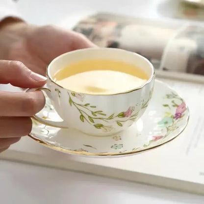A delicate white porcelain teacup with elegant gold flower trim filled with yellow tea in a decorbites saucer set