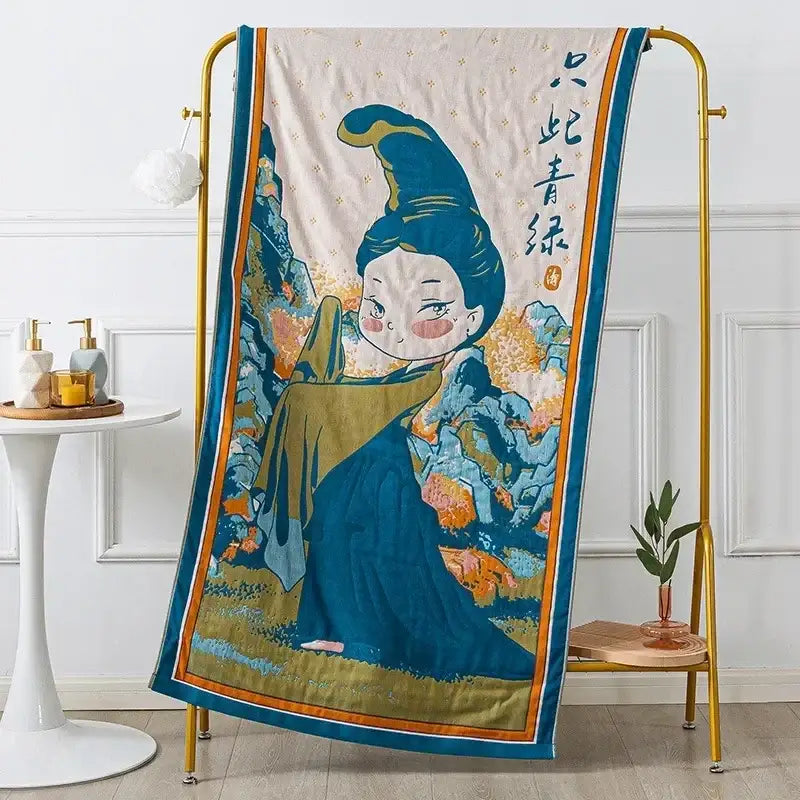 A cream-colored decorbites cotton gauze bath towel with blue and gold embroidered ancient woman design and mountain backdrop