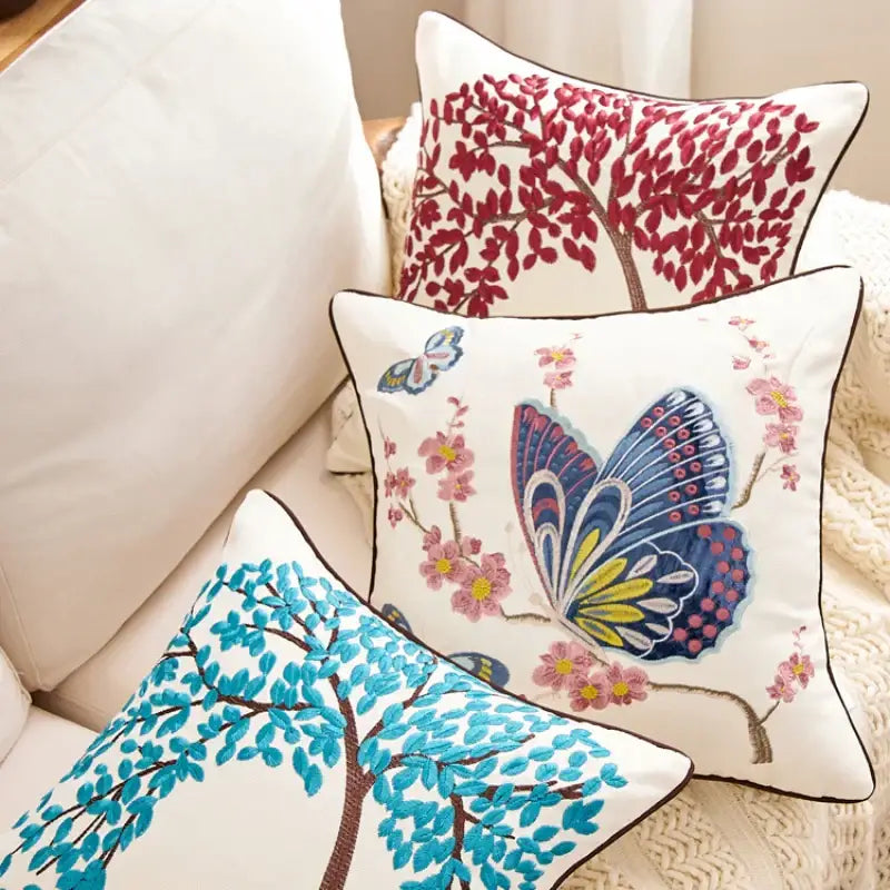 Three embroidered cushion covers with vibrant tree and butterfly designs in red blue and turquoise on cream fabric for sofa