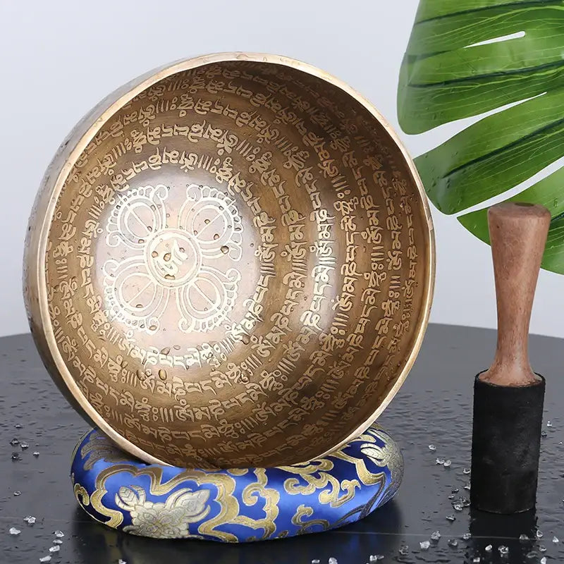 A gold engraved tibetan singing bowl with sanskrit script and floral design resting on an ornate blue cushion