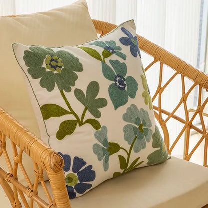 A square farmhouse green and blue floral pillow cover embroidered on cream fabric resting on a wicker chair decorbites decorative farmhouse throw
