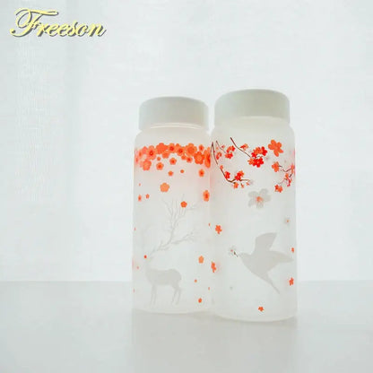 Two frosted glass water bottles with white lids featuring orange cherry blossoms and a deer and red-orange blossoms with a dove design