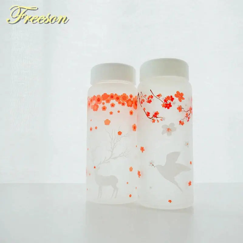 Two frosted glass water bottles with white lids featuring orange cherry blossoms and a deer and red-orange blossoms with a dove design