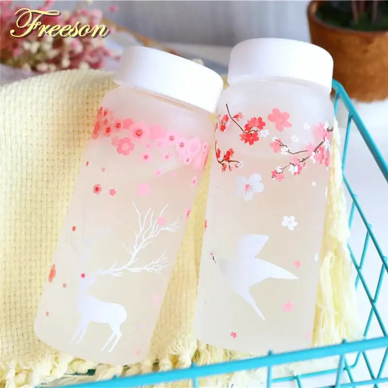 Two frosted glass water bottles with white lids featuring pink cherry blossoms, a deer, and red floral bird designs