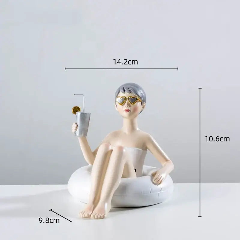 A resin figure sculpture of a woman with short gray hair and gold heart sunglasses sitting in a white inflatable ring holding a cup