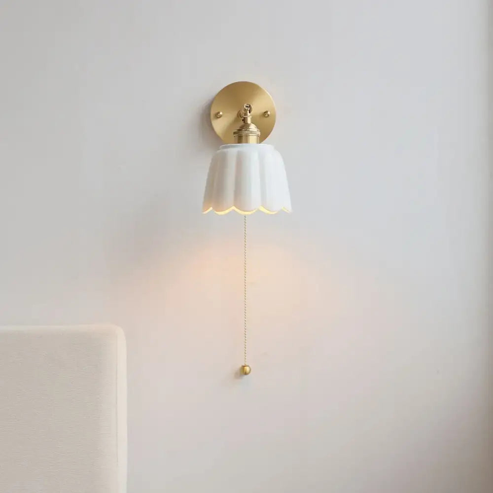 Delicate white ceramic wall light fixture with scalloped edge and gold pull chain for elegant bedroom lighting
