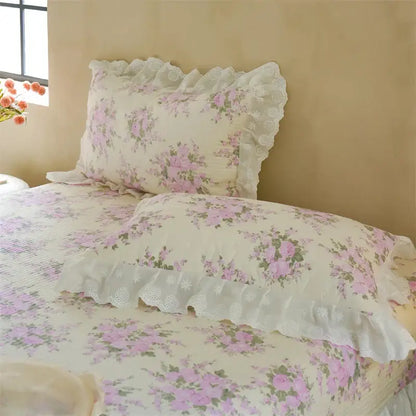 A delicate cream pillow sham with soft pink floral patterns and white lace trim in a quilted bedspread set