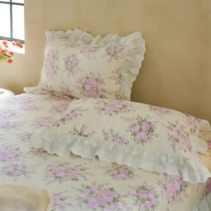 A delicate cream pillow sham with soft pink floral patterns and white lace trim in a quilted bedspread set