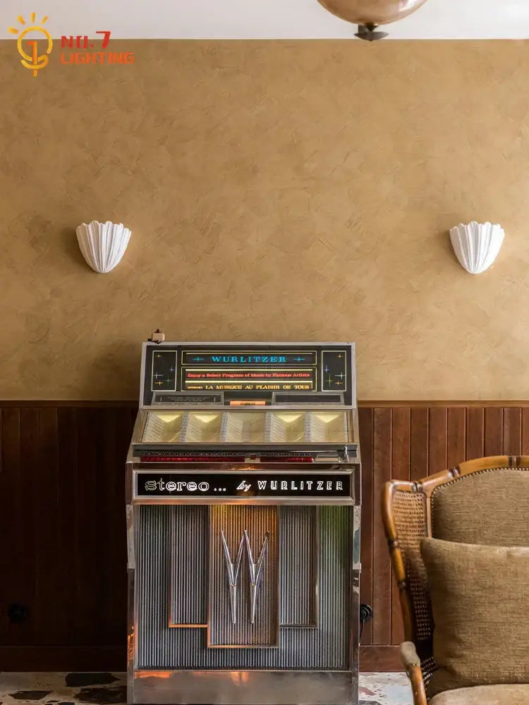 A vintage chrome and black wurlitzer jukebox with blue neon signage next to a french medieval white shell ribbed gypsum wall lamp