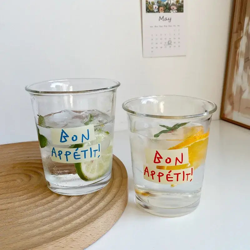 Two clear glass tumbler cups with handwritten bon appétit label filled with ice and citrus slices for kitchen use