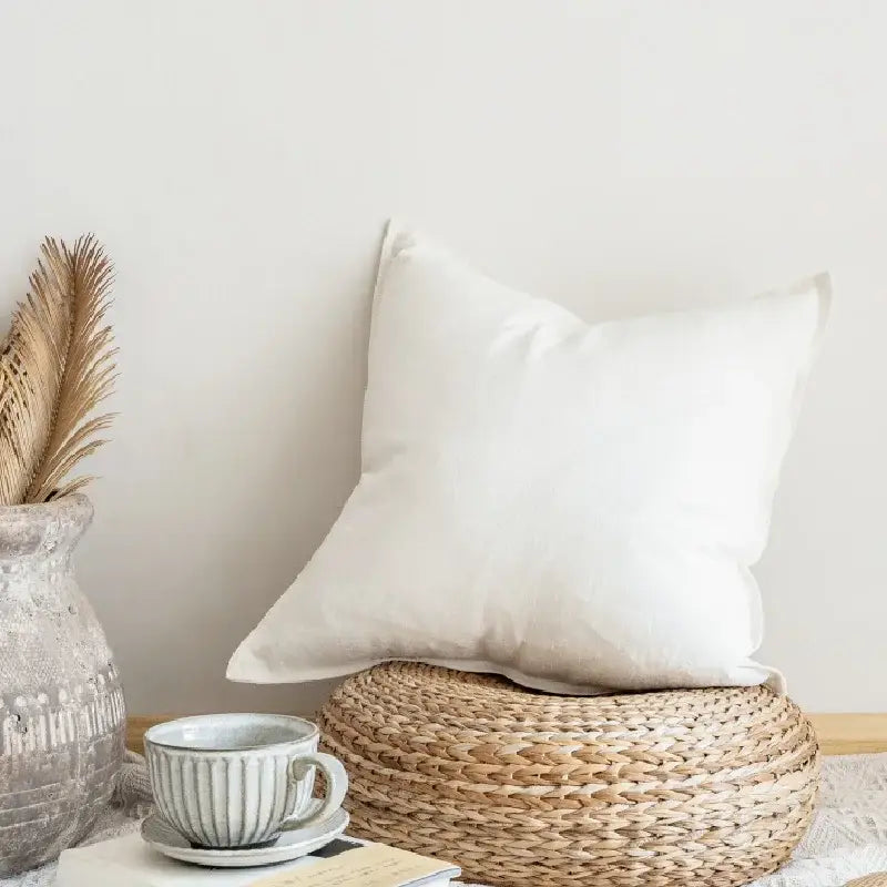 A soft white decorbites linen pillow cover with subtle texture resting on a woven wicker ottoman for living room decor