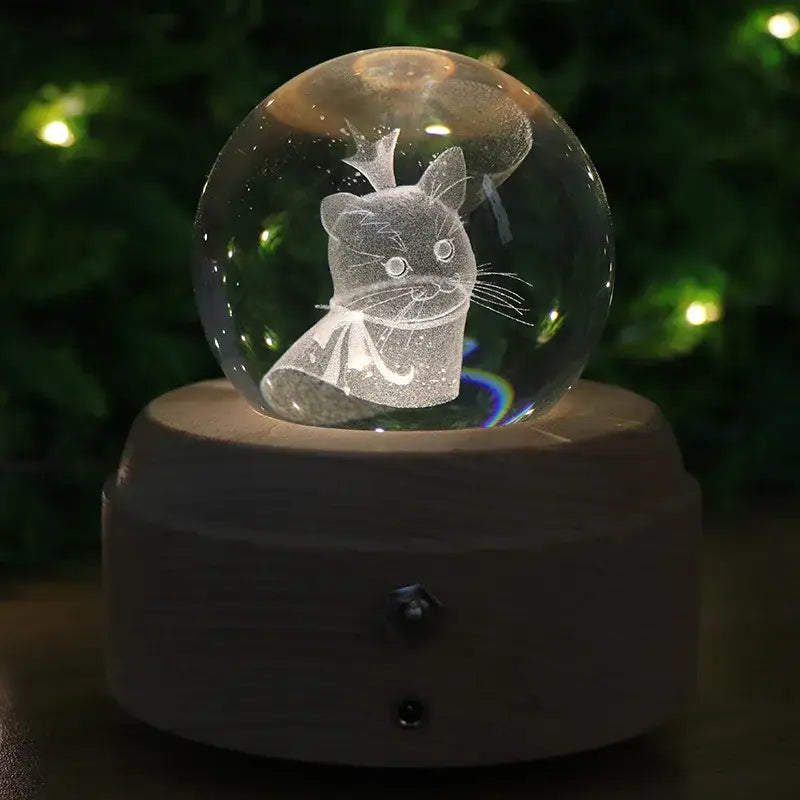 A clear glass crystal ball music box with an engraved black cat inside resting on a wooden base with light glow