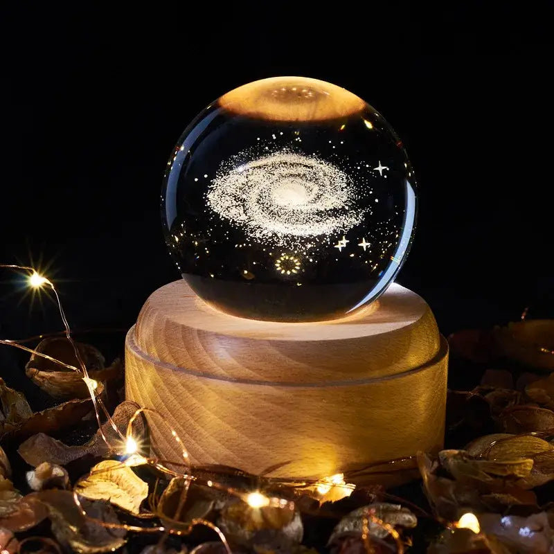 A crystal ball music box with a glowing spiral galaxy design on a clear glass orb resting on a warm wooden base