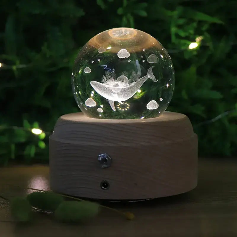 A clear glass crystal ball music box with an engraved white whale and clouds on a wooden rotating base with light glow