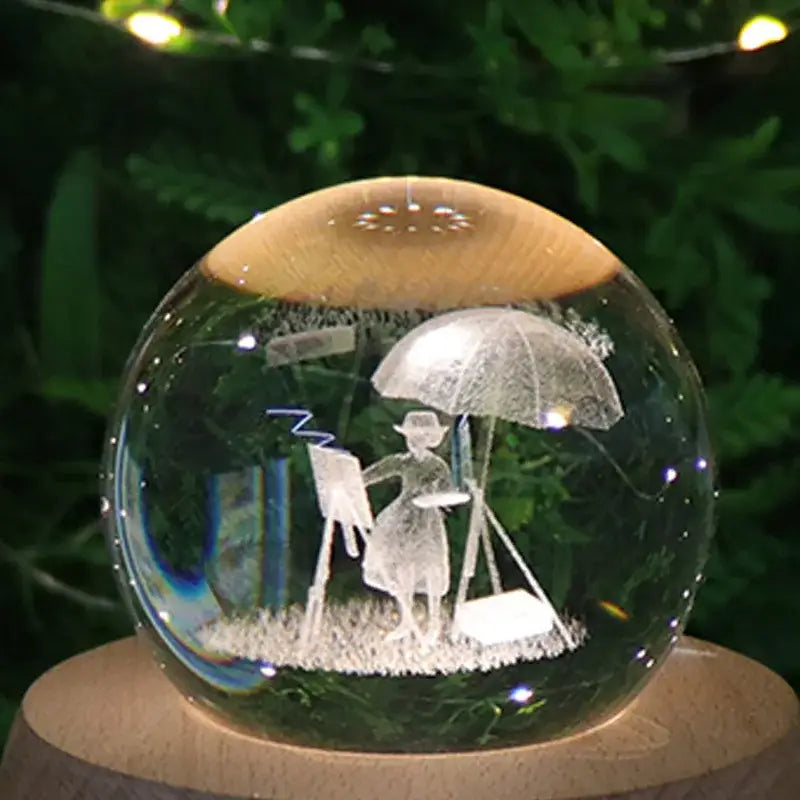 A clear glass crystal ball music box with wooden top and base featuring an engraved painter scene and rotating light glow