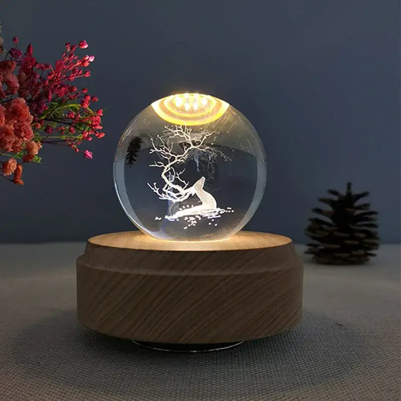 A clear glass crystal ball music box with an etched white deer design and warm yellow light on a wooden base