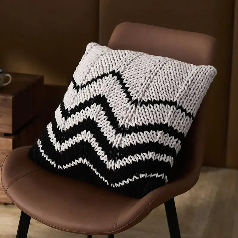 A black and white chunky knit geo knit stripe square pillow with a bold zigzag pattern on a brown leather chair