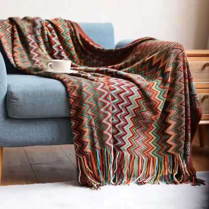 A cozy multicolored knit geometric aztec baja blanket in mustard and blue draped over a blue armchair with fringed edges
