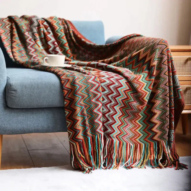 A cozy multicolored knit geometric aztec baja blanket in mustard and blue draped over a blue armchair with fringed edges