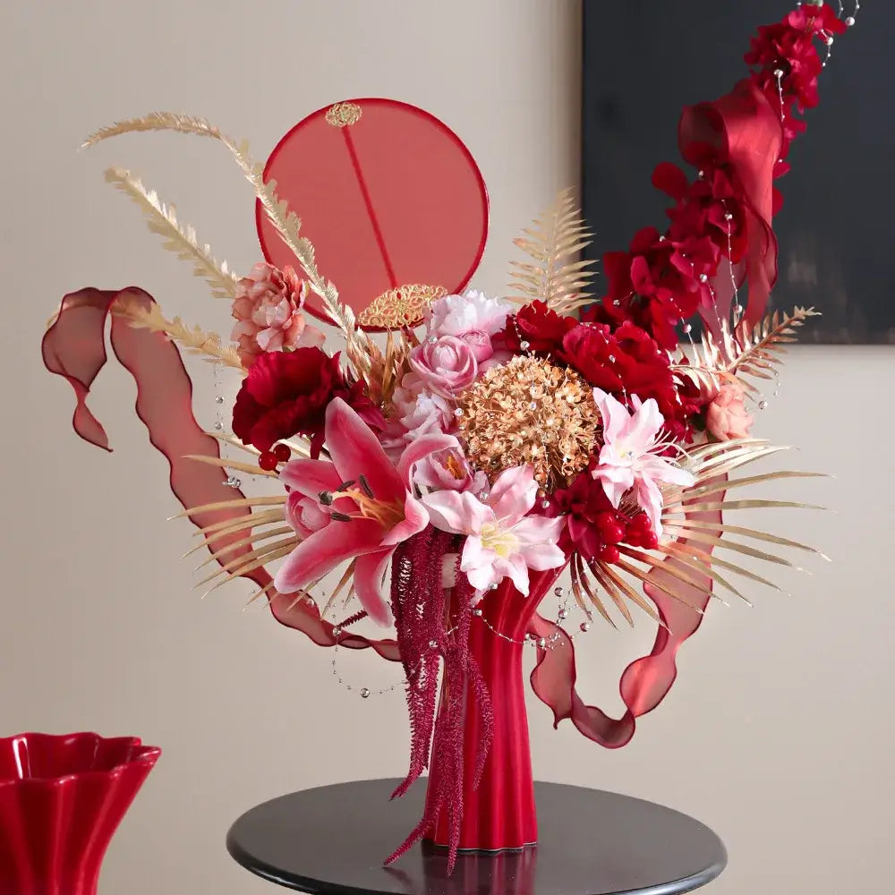 A vibrant red ceramic vase with a geometric fold pattern holds an elaborate floral arrangement with gold accents