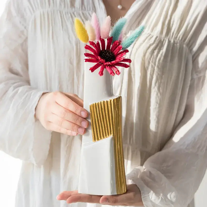 White ceramic vase with geometric gold striped ribbed accent holding red gerbera daisy and colorful feathers for living room hydroponics