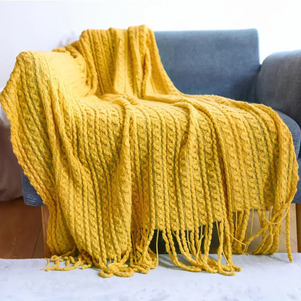 A cozy mustard-yellow cable knit sofa blanket with fringed edges draped over a gray armchair decorbites geometric knit