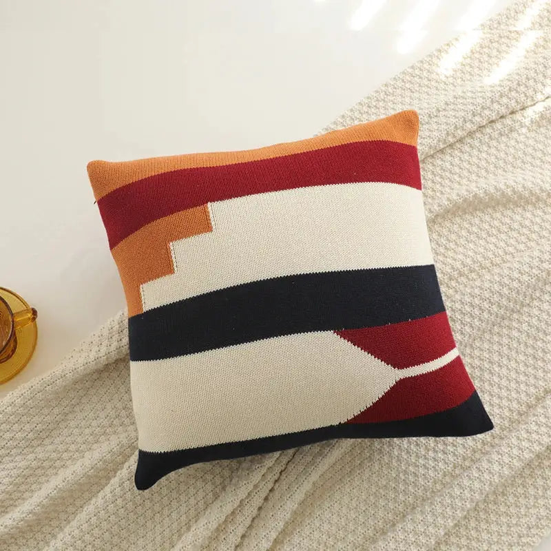 A square knitted cushion cover featuring bold orange red cream and black stripes with a textured knit pattern