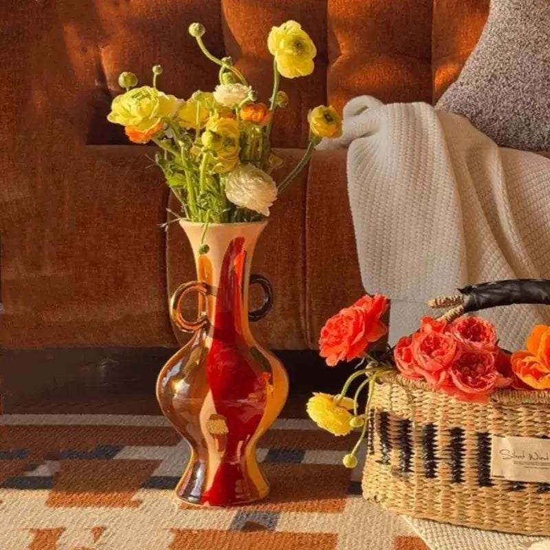 Vibrant red and amber glass vase with dual handles holding yellow and white ranunculus flowers in living room decor with geometric horse head