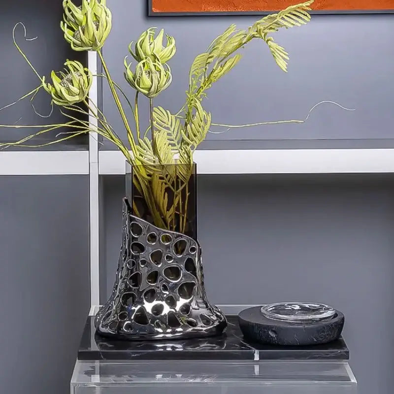 Black metallic glass vase with perforated organic design holding green artificial flowers for geometric acrylic decorbites