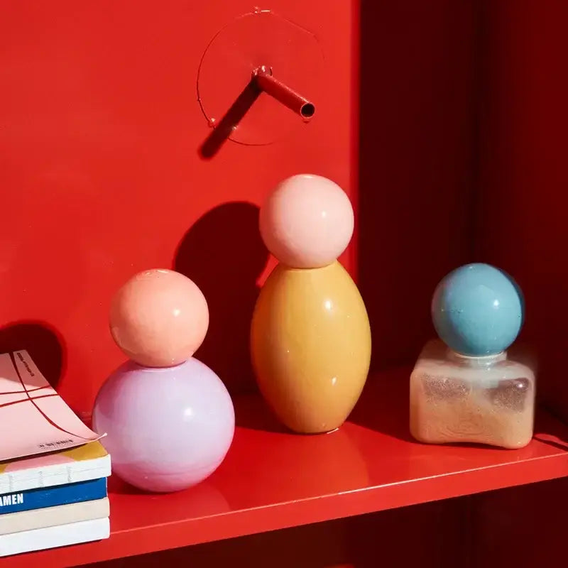 Pastel-colored ceramic figurines with smooth forms displayed beside a geometric sphere glass vase with lid for bedroom storage