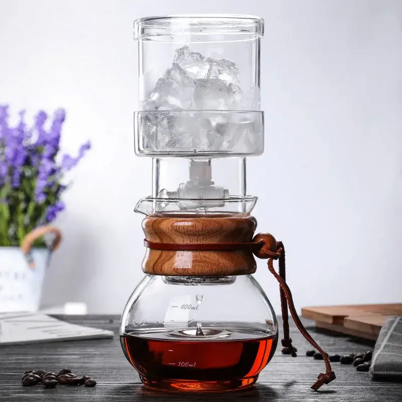 Glass cold brew coffee maker with clear glass, wooden lid, brown leather strap, and top chamber filled with ice cubes