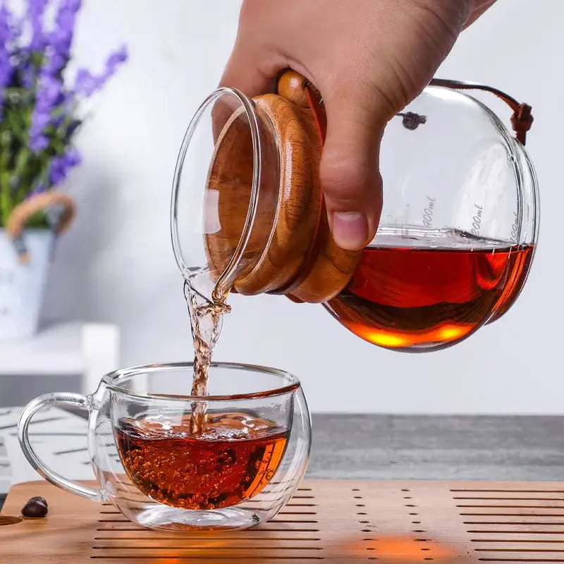 A clear glass cold brew coffee maker with wooden lid and handle pours amber brew coffee into a double-walled glass cup