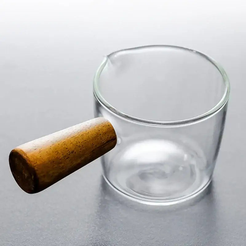 Clear glass espresso measuring cup with a smooth light brown wood handle for kitchen milk jug use