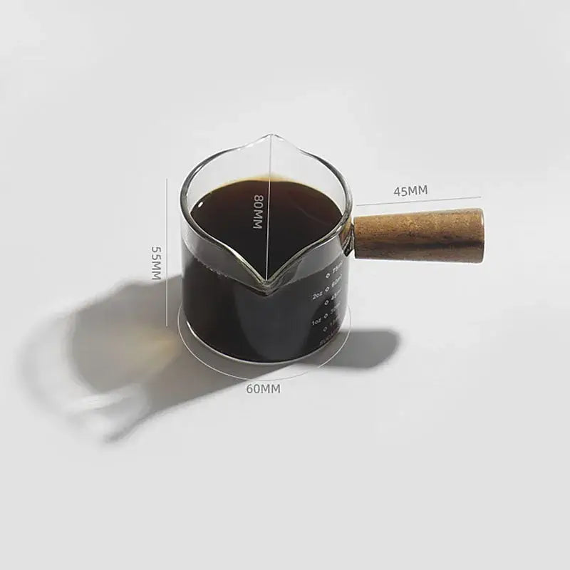 Clear glass espresso pitcher with wooden handle filled with dark liquid for precise kitchen measuring use