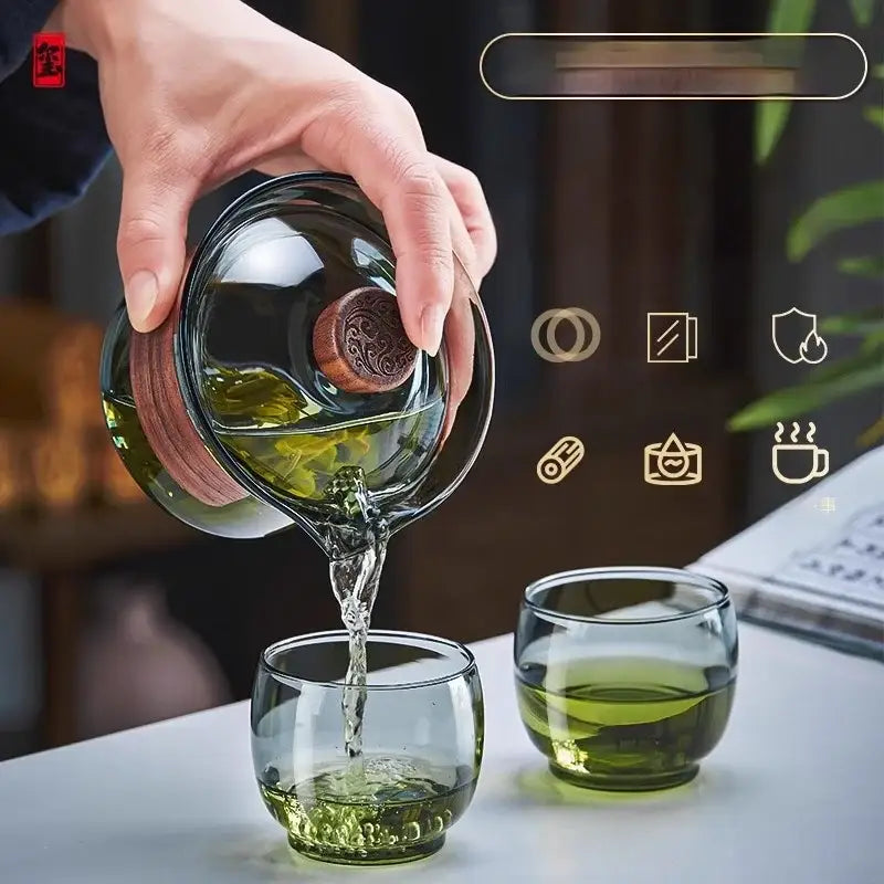 A clear glass gaiwan cup set with wooden lid and handle pouring green tea into matching glass cups