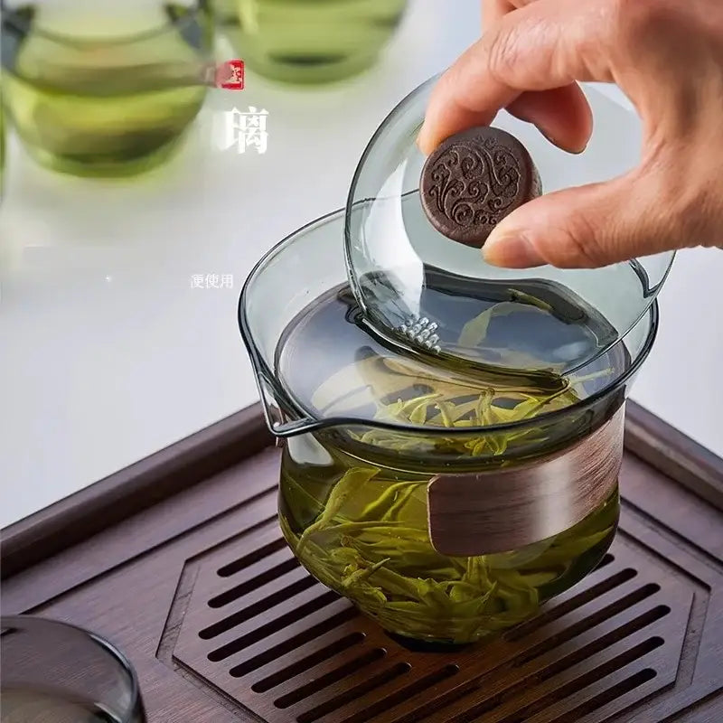 A dark brown engraved tea cake lifted from a clear glass gaiwan cup set with wood ring tea filter and green tea leaves