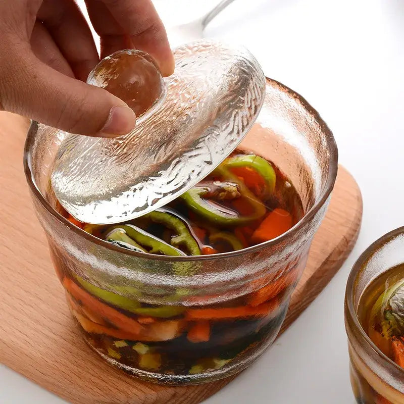 Clear glass kimchi storage jar with textured lid filled with colorful pickled vegetables in dark brine by decorbites