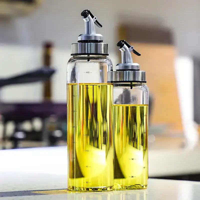 Two transparent glass seasoning dispenser bottles with stainless spouts and black nozzles filled with golden liquid