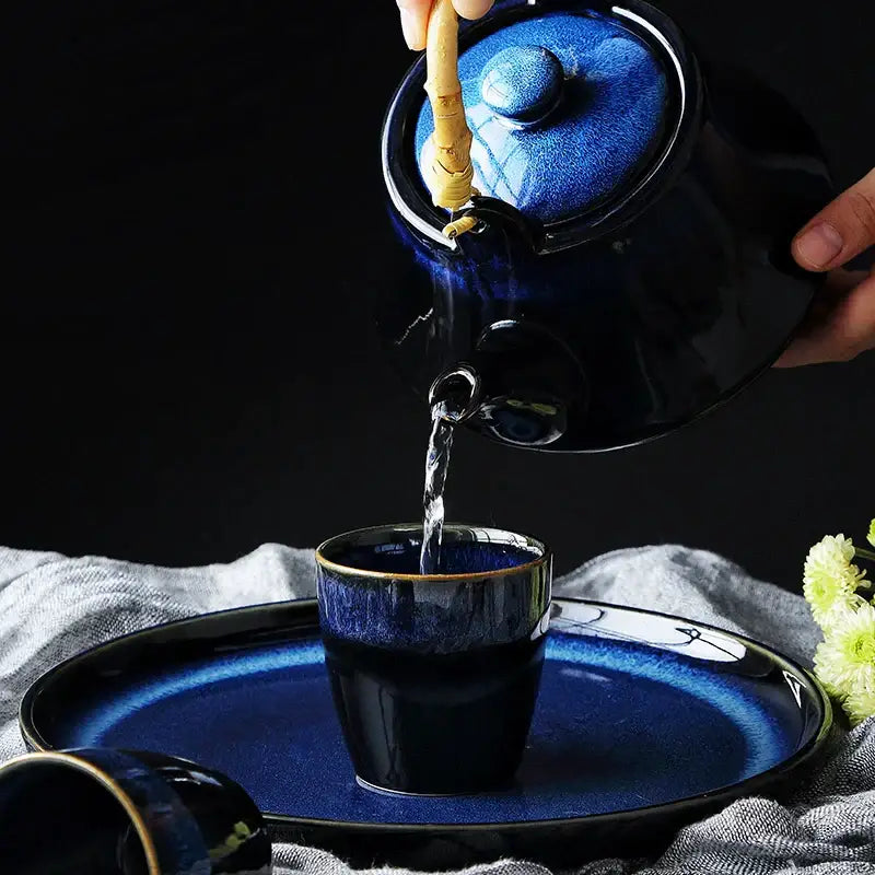 Glossy deep blue ceramic tea kettle with gold rim and bamboo handle pouring into matching cup on coordinating plate decorbites blue ceramic