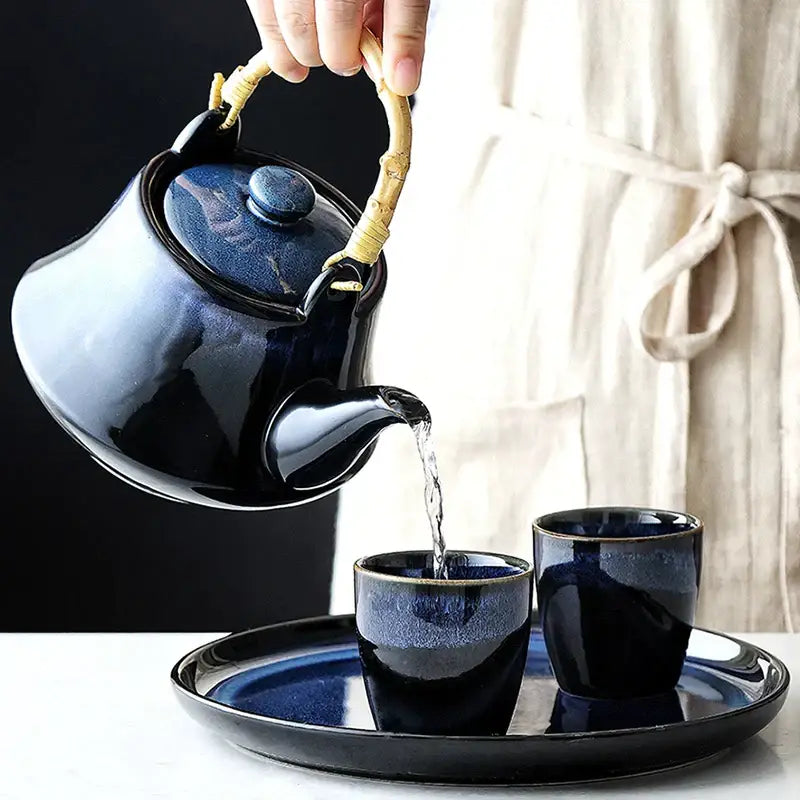 A glossy dark blue ceramic tea kettle with bamboo handle pouring tea into matching cups on a decorbites tray
